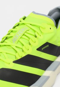 Zapatilla deportiva verde lima con detalles en negro, que cuenta con una parte superior de malla, un sistema de cordones distintivo y una suela de perfil bajo. Marca: Adizero.