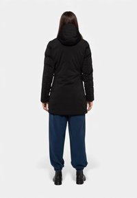 Cappotto imbottito nero con cappuccio, caratterizzato da una silhouette lunga. Indossato sopra pantaloni blu larghi e stivaletti neri alla caviglia.