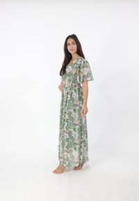 Maxi jurk van doorschijnende groene stof met bloemenpatronen, V-hals, elastische taille, korte vlindermouwen en doorzichtige hakken.