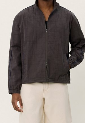 Un homme porte une veste zippée gris foncé avec un motif discret en quadrillage et un short couleur crème, debout devant un fond clair uni.