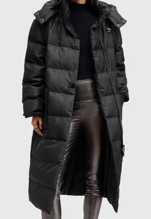 Personne portant un long manteau matelassé noir par-dessus un haut noir et des leggings métalliques argentés brillants, les mains détendues le long du corps.