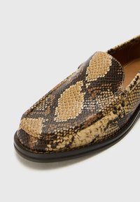 Des chaussures slip-on marron et beige avec un motif de cuir de serpent texturé, dotées d'un bout rond et d'une semelle noire basse.