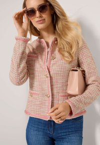 Rosa-weiße Tweedjacke mit goldenen Knöpfen, gewellten Kanten und zwei Vorderaschen. Kombiniert mit einer hellrosa Handtasche und übergroßen Sonnenbrillen.