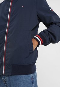 Muž v námořnickém modrém saku s červeným, bílým a černým pruhem na zipu a na manžetách, s rukou v kapse, kombinovaný s modrými džíny.