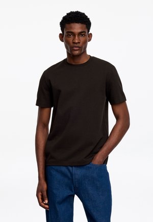 Jeune homme aux cheveux courts et bouclés portant un t-shirt noir uni à manches courtes et un jean bleu, debout avec une main dans la poche.