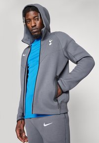 Grå zip-up hoodie med hætte, der har et logo på brystet og to sidelommer. Båret over en blå atletisk top og grå bukser med et logo.