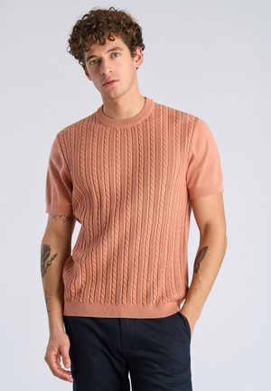 Junger Mann mit lockigem Haar, der einen strukturierten, kurzärmligen Pullover in Lachsfarbe und eine dunkle Hose trägt, mit einer Hand in der Hosentasche stehend.