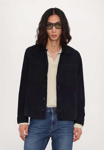 Veste bleu marine foncé avec deux poches poitrine, fermeture à boutons et manches longues. Portée sur une chemise beige clair et associée à un jean bleu.