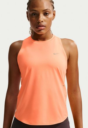 Młoda kobieta z zaplecionymi włosami, ubrana w pomarańczowy sportowy top Nike bez rękawów na jasnym, jednolitym tle.