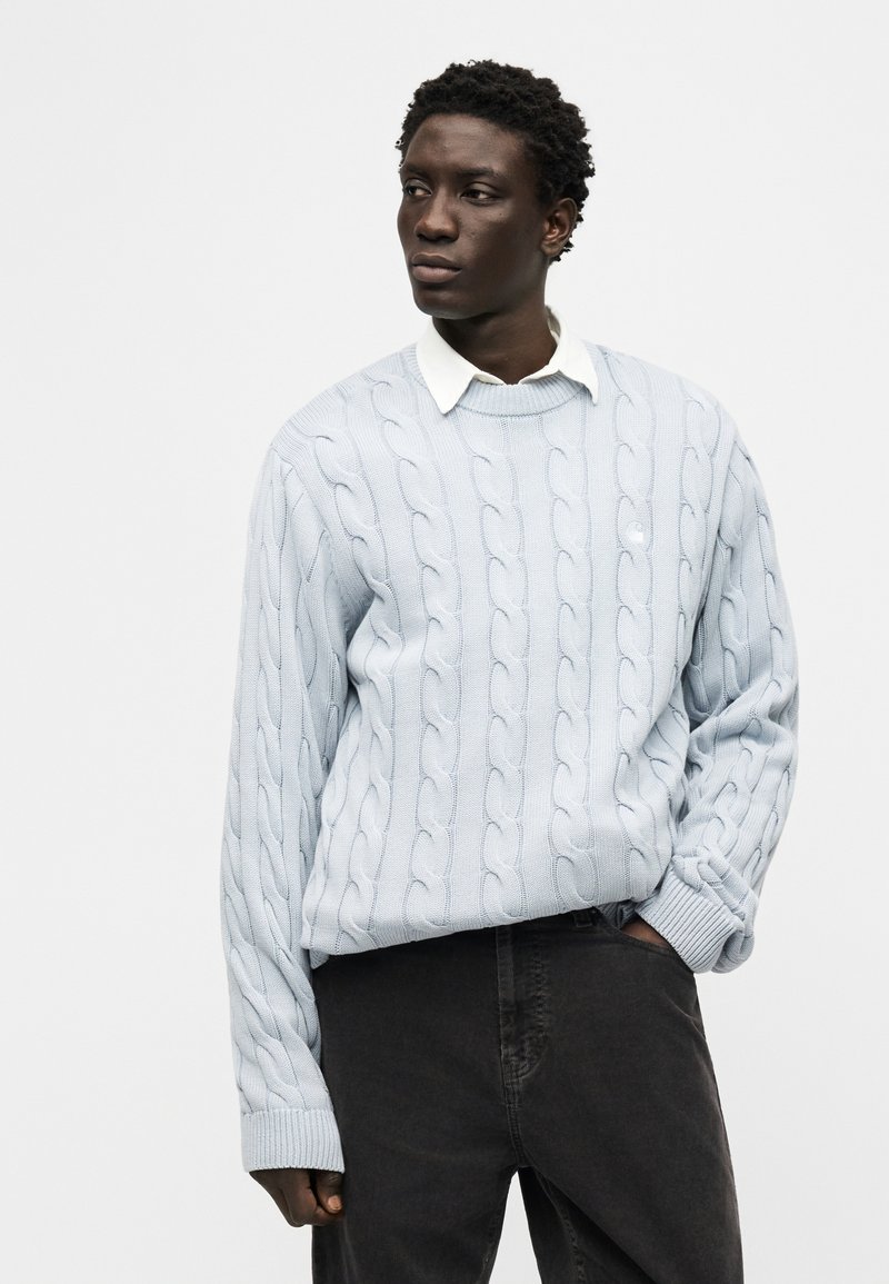 Homme en pull bleu clair à torsades par-dessus une chemise blanche à col, main dans la poche, regardant sur le côté sur un fond uni.