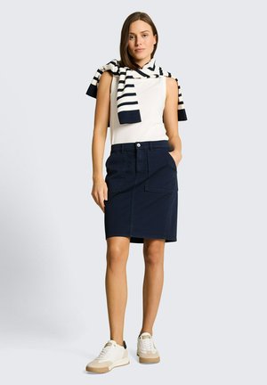 Femme portant un top blanc sans manches, une jupe bleu marine longueur genou avec des poches avant, un pull rayé bleu marine et blanc drapé sur les épaules, des baskets blanches.