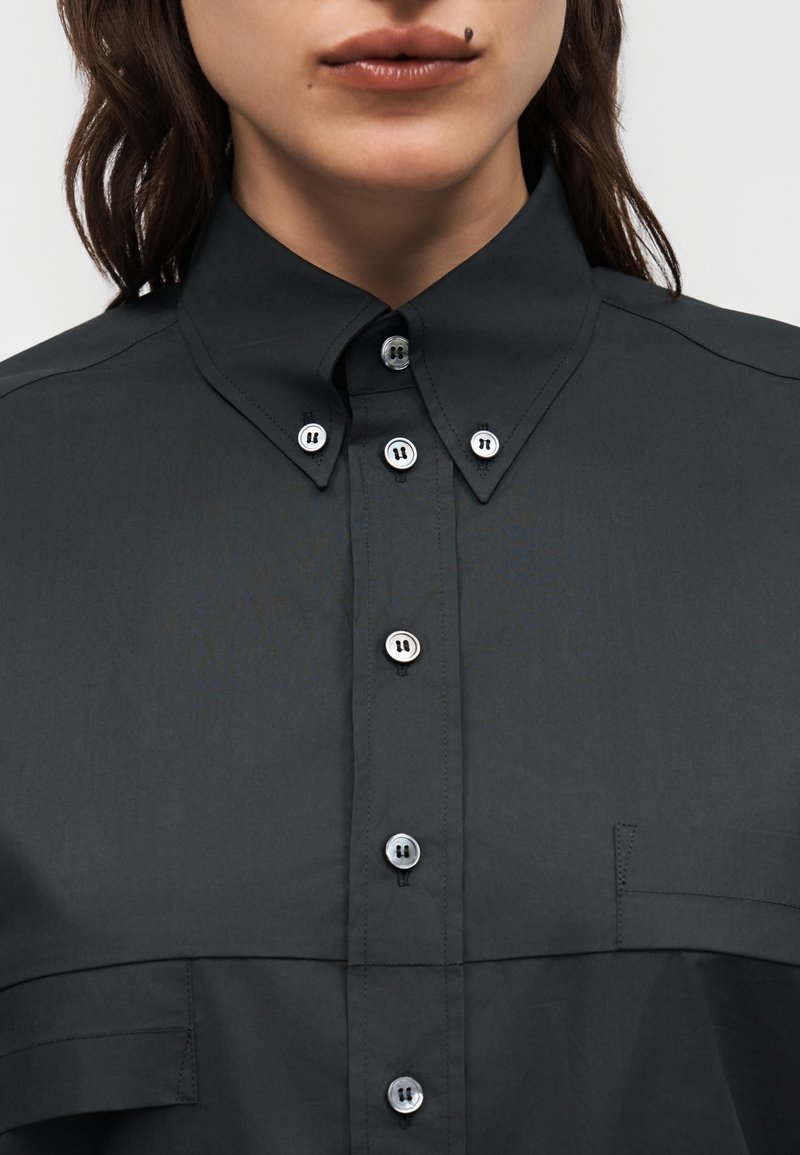 Gros plan d'une personne portant une chemise foncée à boutons blancs et col pointu, montrant les lèvres et un grain de beauté au-dessus de la lèvre supérieure.