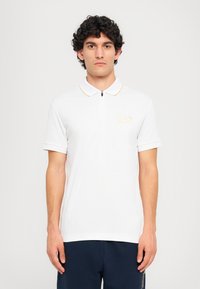 Balta polo marškiniai su užtrauktuko apykakle, trumpomis rankovėmis ir geltonu akcentu aplink apykaklę. Rodyklėje yra logotipas ant krūtinės. Paprastas dizainas.