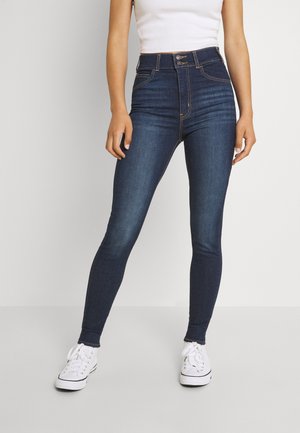 Femme portant un jean skinny bleu marine taille haute et des baskets blanches, debout devant un fond uni.