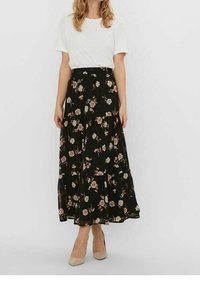 Vrouw in een witte blouse met korte mouwen, zwarte bloemen midi-rok en beige hakken, staande tegen een plain witte achtergrond.