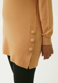 Langærmet, ribbet tunika i blød, ferskenfarvet strik. Har tre strukturerede knapper langs siden, med en rundet kant og sidesplitter.