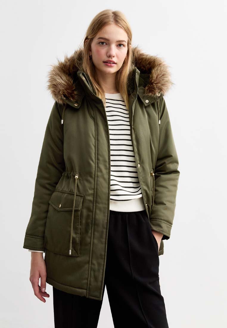Parka vert olive avec une capuche en fausse fourrure, fermeture à glissière et cordon de serrage, deux poches avant et un ourlet côtelé, portée sur un haut rayé.