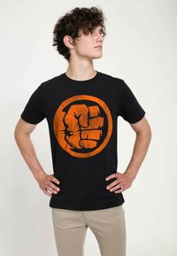 T-shirt nera in cotone con una grafica di un pugno arancione scolorito all'interno di un cerchio. Modello che indossa pantaloni color beige chiaro, in piedi con le mani sui fianchi.