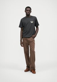 Melns kokvilnas T-krekls ar baltu un dzeltenu grafisko logotipu, apvienots ar brūnām platām biksēm un brūniem laivu apaviem, stāvot pret neitrālu fonu.