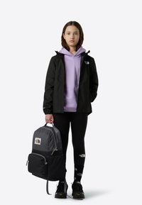 Must veekindel jope, lavendli tooniga kapuutsiga pusa, mustad retuusid, halli ja musta värvusega seljakott, mustad jalanõud kollaste detailidega, nähtavad logod.