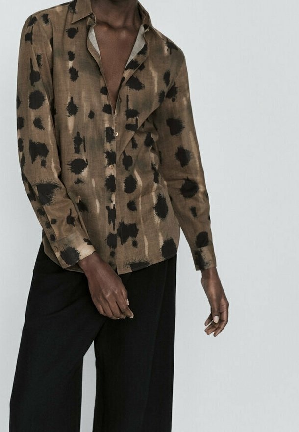 Ruskea kuvioitu paita, jossa mustia abstrakteja pilkkuja, pitkät hihat, lyhyt kaulus ja nappikiinnitys. Yhdistetty mustiin leveälahkeisiin housuihin.