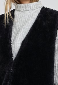 Černá vesta z umělé kožešiny přes světle šedý žebrovaný svetr s rolákem. Vesta má šálový límec a měkký, texturovaný povrch.