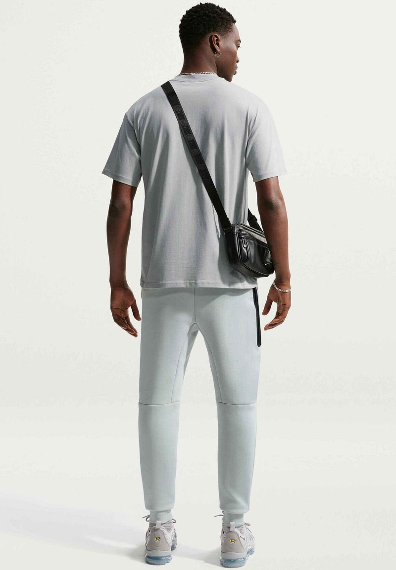 Mann steht mit dem Rücken zur Kamera, trägt ein hellgraues T-Shirt, passende Jogginghosen, weiße Sneakers und eine schwarze Umhängetasche.