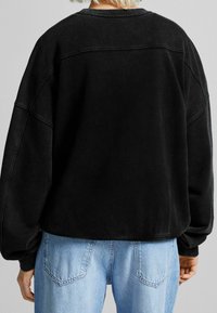 Sudadera negra de gran tamaño con cuello y puños acanalados. Textura suave, dobladillo curvado y detalles de costura visibles. Combinada con unos vaqueros de color azul claro.