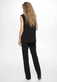 Femme avec de longs cheveux blonds ondulés portant un haut noir sans manches et un pantalon noir, se tenant de dos à la caméra sur un fond blanc.