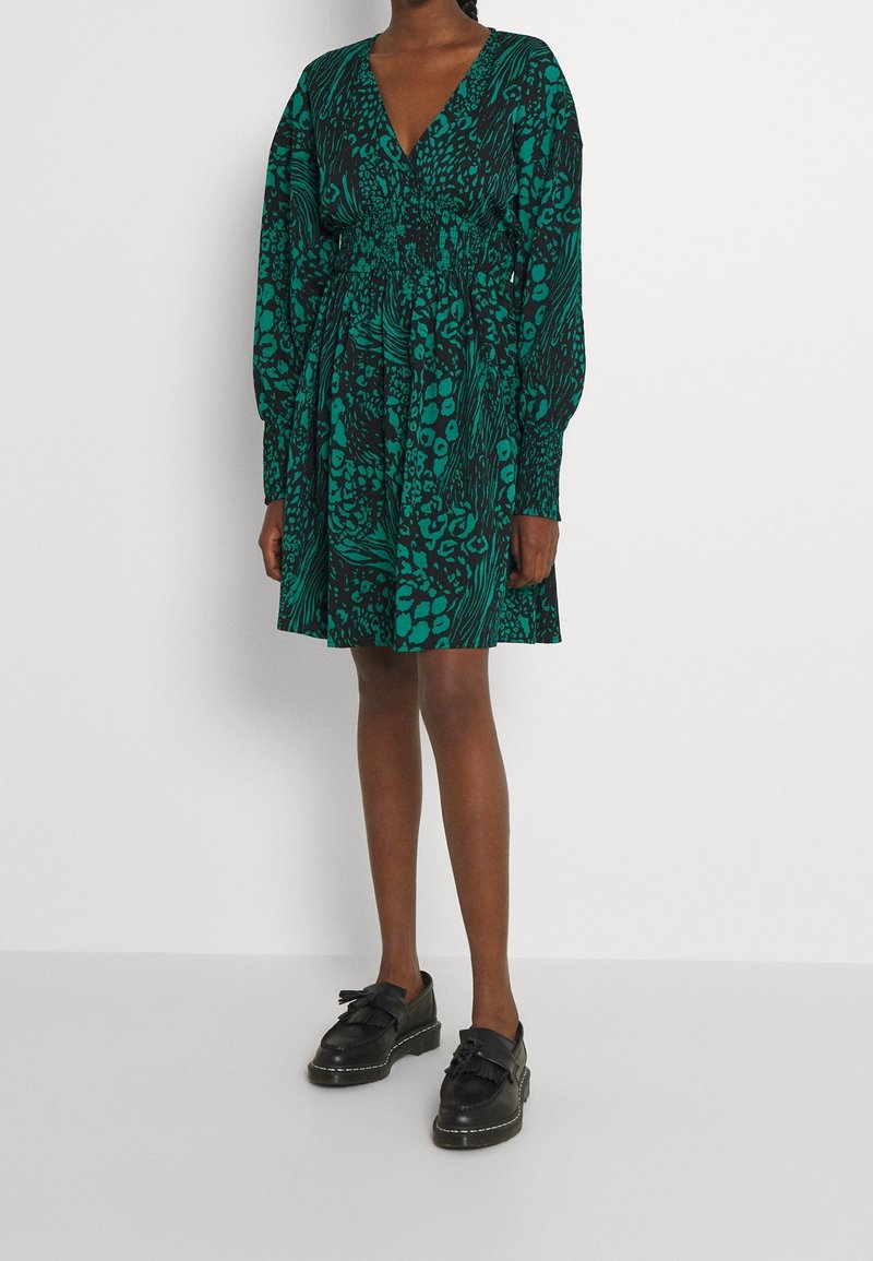 Femme portant une robe à imprimé animal vert et noir à manches longues et des mocassins noirs, debout sur un fond blanc.