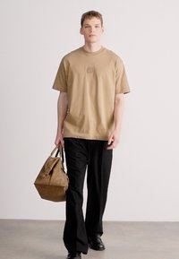 Καφέ βαμβακερό t-shirt με μικρό κεντημένο λογότυπο, συνδυασμένο με μαύρο παντελόνι με φαρδιά πόδια. Το μοντέλο κρατά καφέ δερμάτινη τσάντα.