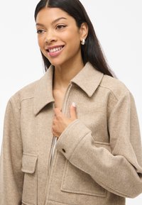 Giacca in misto lana beige con zip frontale, grande colletto e tasche frontali. Presenta una vestibilità rilassata, cuciture minime e una texture liscia.