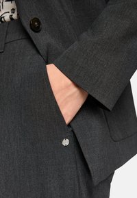 Dark gray tailored blazer with a smooth texture, featuring a front pocket, a button closure, and subtle stitching details.