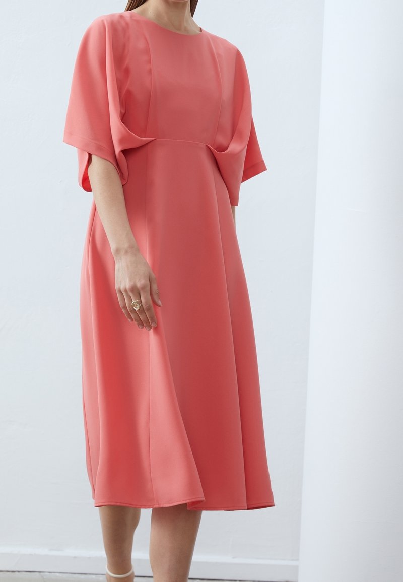 Femme portant une robe midi rose corail avec de larges manches drapées, debout contre un mur blanc.