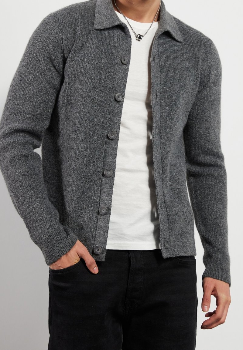 Cardigan grigio lavorato a maglia con scollo con colletto, chiusura frontale con bottoni e polsini a coste, indossato sopra una maglietta bianca. Pantaloni neri visibili.