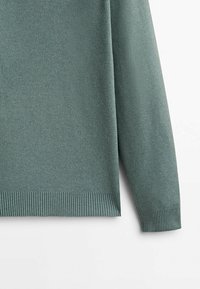 Manche et ourlet d'un pull en maille vert clair avec poignets et ceinture côtelés sur fond blanc.