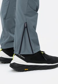 Pantalons d'extérieur gris avec zips sur les côtés, poignets élastiques, associés à des chaussures noires avec un maillage texturé et un accent jaune sur la semelle.