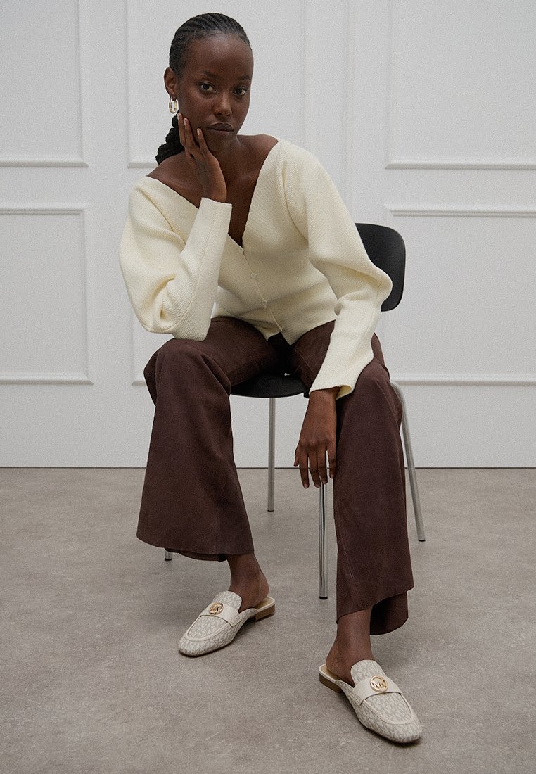 Romige gebreide cardigan, bruine wijde broek en lichtkleurige loafers met een textuurpatroon en decoratieve hardware. Zit op een stoel.