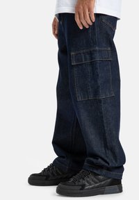 Modré denimové cargo nohavice s voľným strihom, bočnými vreckami a viditeľným oranžovým prešívaním. Skombinované s čiernymi nízkymi teniskami.