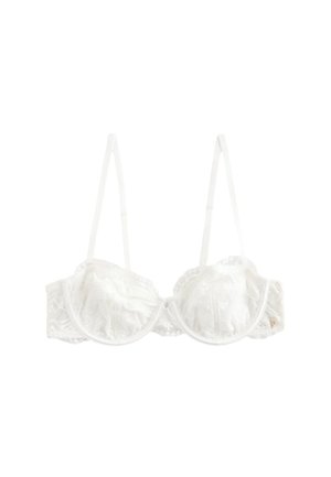 Vrouw met krullend donker haar die een crèmekleurige kanten bh en kanten slip met hoge taille draagt, staande binnen bij een tafel en doorschijnende gordijnen.