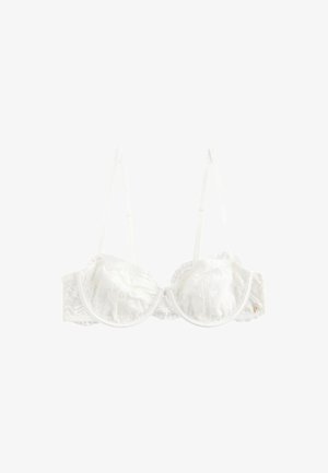 Vrouw met krullend donker haar die een crèmekleurige kanten bh en kanten slip met hoge taille draagt, staande binnen bij een tafel en doorschijnende gordijnen.
