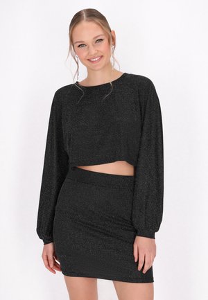 Mladá žena se usmívá, má na sobě černý crop top s dlouhým rukávem a laděnou úzkou minisukni s třpytivým detailem, stojící na bílém pozadí.
