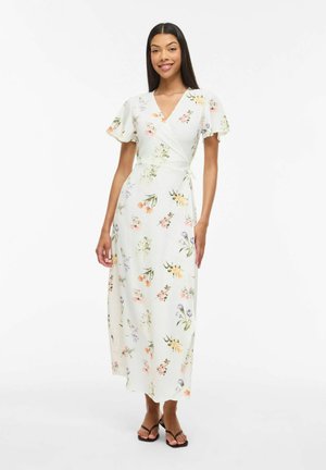 Žena v bílé zavinovací šaty s krátkými volánovými rukávy a vícebarevným květinovým potiskem, stojící a usměvající se na jednobarevném pozadí.