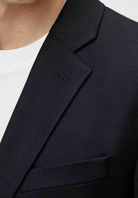 Granatowa marynarka teksturowana z klapami w kształcie litery notched, pojedynczą kieszenią na piersi oraz subtelnymi detalami szycia. Nosi ją się na białej koszuli.