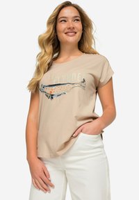 Camiseta de algodón beige con un diseño de pez impreso en azul y naranja. Con mangas cortas y un corte relajado. Combinada con pantalones blancos.