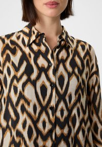 Een button-up shirt met een luipaardprint in zwart, tan en crème. Gemaakt van lichtgewicht stof met een zachte textuur.