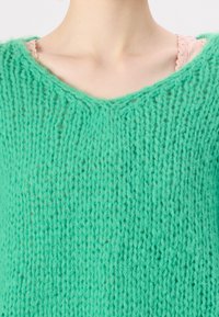 Mintgrøn strikket sweater med V-hals, der har et tekstureret, løst vævet stof. Laget over en lys pink top.