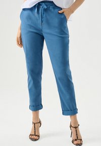 Pantalons légers et fuselés bleus avec une taille à cordon, des poignets roulés et une texture lisse, associés à des sandales à talons hauts à lanières.