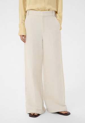 Kvinde iført højttaljede, brede ben i tekstureret cremefarvet bukser, kombineret med en lysegul langærmet skjorte og brune sandaletter med åben tå.