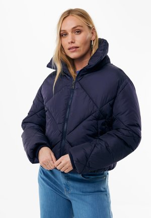 Frau mit langen blonden Haaren, die eine dunkelblaue Steppjacke mit Reißverschluss und hellblaue Jeans vor weißem Hintergrund trägt.